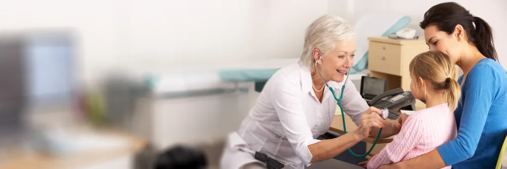 This image shows a doctor's surgery examination room where a senior doctor is examining a child with a stethascope, while an adult holds her for support